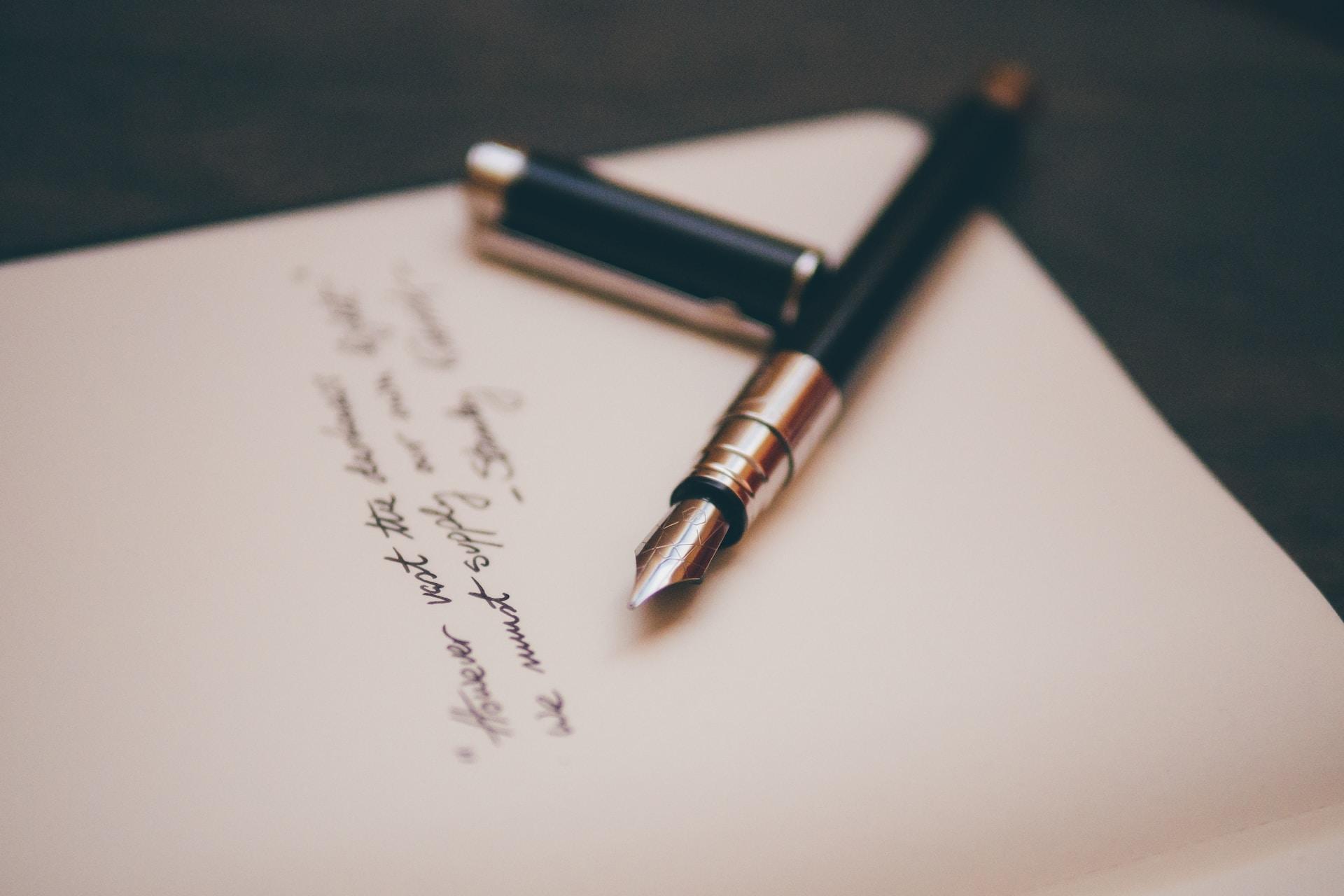 Calligraphy pen and pen lid on top of a notebook containing white pages and non-discernable black cursive writing on top of a dark-colored table.